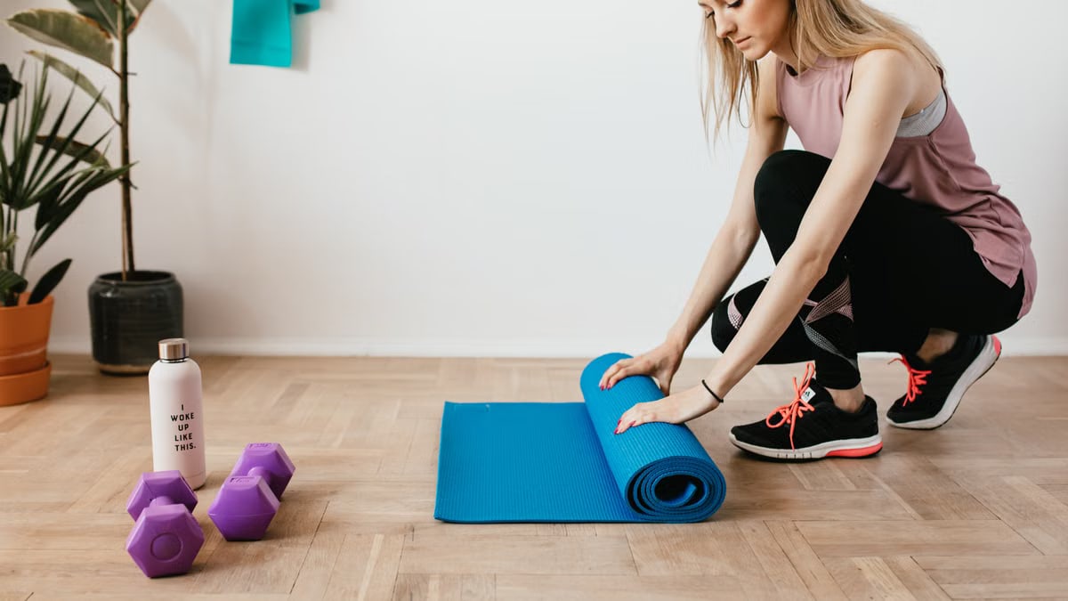 Side view of young female athlete in sportswear unfolding blue sport mat and looking down while sitting on floor near dumbbells and water bottle preparing for training indoors